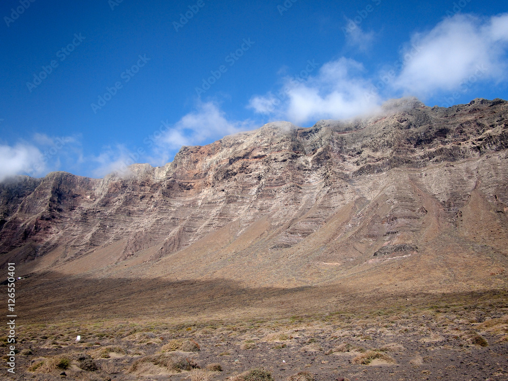 White clouds descend from the slopes of volcanic mountains to the valley on a background of deep blue sky. Famara, Lanzarote, Canary Islands, Spain
