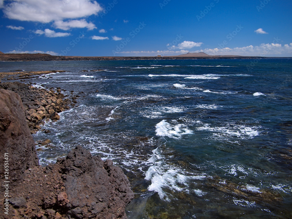 Obraz premium Ocean waves breaking on the rocky coast of hardened lava with caverns and cavities. Deep blue sky with white clouds and mountains and volcanoes on the horizon. Lanzarote, Canary Islands, Spain