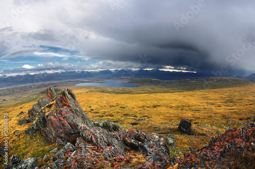 Colorful rock in the background of highland steppe lake with snow covered mountains under a stormy cloudy dramatic blue sky Plateau Ukok, Altai, Siberia, Russia