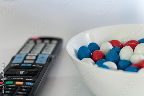 White bowl with red, white and blue candy dragee and tv remote on white table