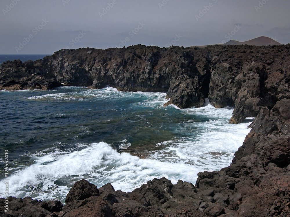 Ocean waves breaking on the rocky coast of hardened lava with caverns and cavities. Mountains and volcanoes on the horizon. Lanzarote, Canary Islands, Spain