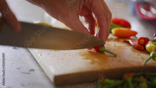 Chef cuts off peduncle from fresh green, yellow, red and orange chili peppers using sharp knife; close-up; 
