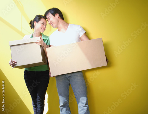 Couple carrying boxes, standing against yellow wall © Alexander