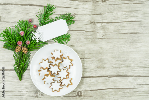Kleiner runder weißer Teller mit Weihnachtsplätzchen, Zimtsternen, einem Tannenzweig und gefrorenen roten Beeren und einem weißen Schild, das beschriftet werden kann, Untergrund Holz, Textfreiraum
