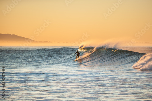 Morning sun glows off surfer as east winds blow tops of waves at Ventura beach.