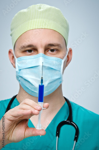The doctor in a green hat and a blue mask holding a syringe with medicine.