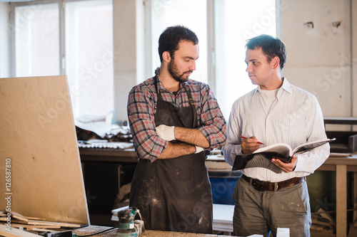 Happy male carpenter showing something to coworker at his notebook papers in workshop