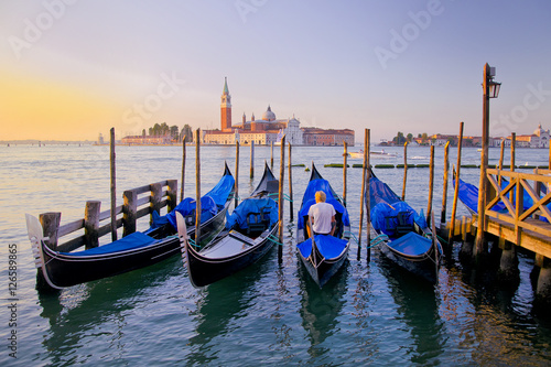 Young man in white clothes sitting in front of Grand Canal in Venice with gondolas against San Giorgio Maggiore church in Italy. Scenery from summer vacation with beautiful colorful morning light.