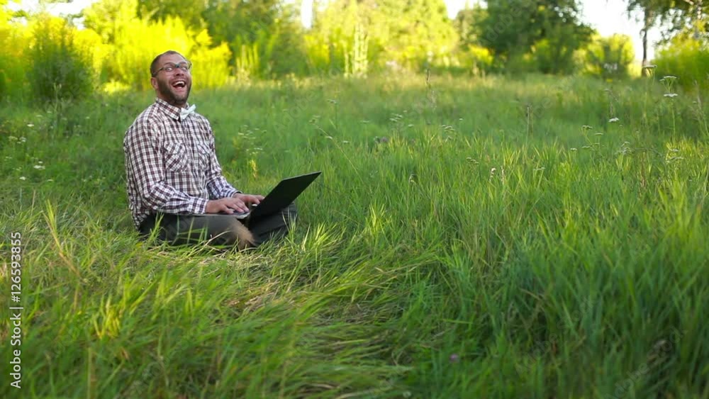 Happy young man in glasses sitting on the meadow and typing on t Happy young man in glasses sitting on the meadow and typing on t