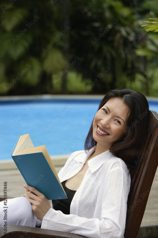 Obraz premium Woman sitting by swimming pool, holding a book, smiling at camera