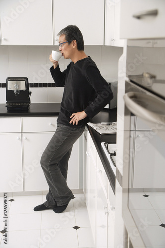 Obraz na płótnie Man in kitchen, leaning on counter, drinking coffee, side profile to camera. w salonie