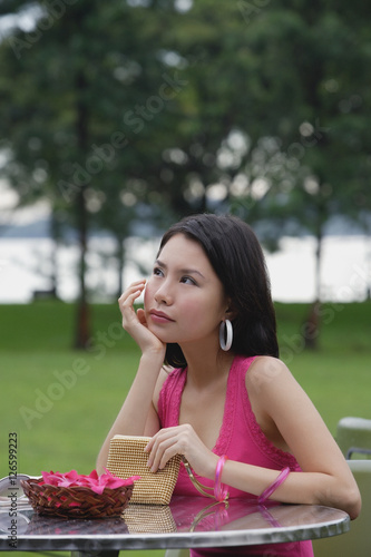 Young woman sitting at outdoor table, hand on chin, looking away © Alexander