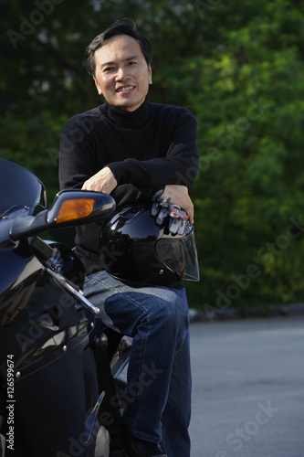 Man leaning on motorcycle, holding helmet, smiling at camera