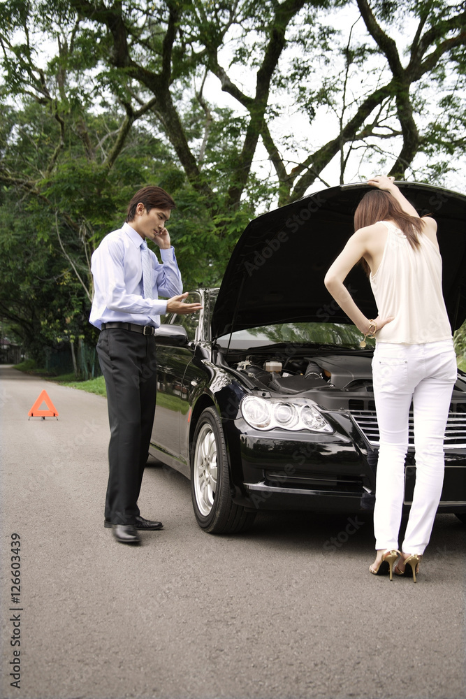 Woman looking under hood of car while man talks on phone