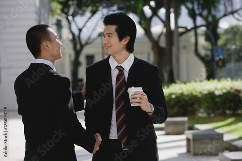 Two men wearing suits greet each other and shake hands in the park