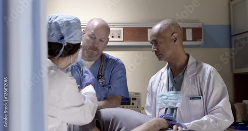 Male and female doctors work with male nurse to care for a male patient with a head injury in hospital examining room.  Recorded hand-held.