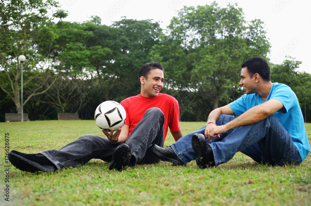 Two men sitting on grass, talking, one holding soccer ball Two men sitting on grass, talking, one holding soccer ball