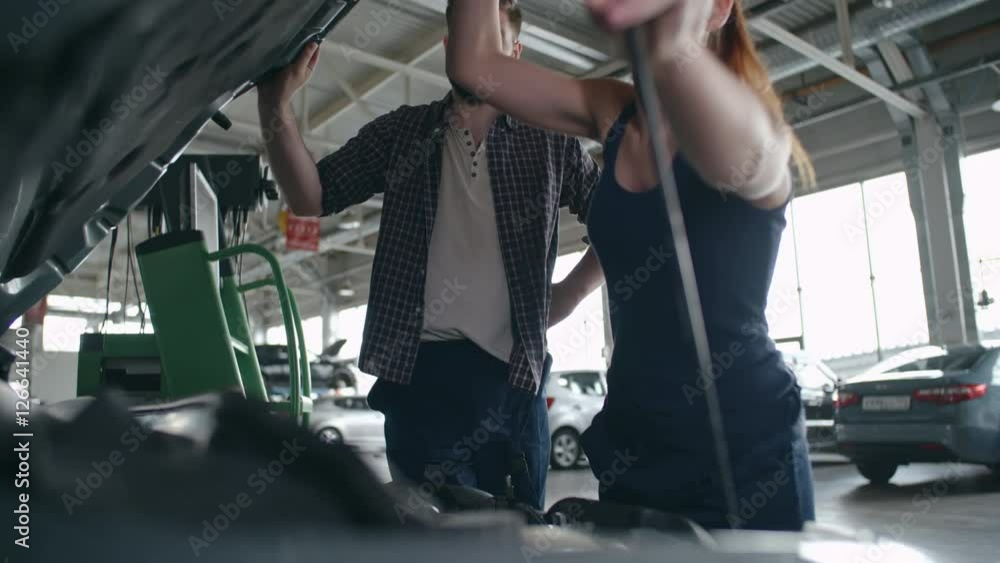 Man and woman in mechanic uniform opening car hood, checking under it and discussing something Man and woman in mechanic uniform opening car hood, checking under it and discussing something