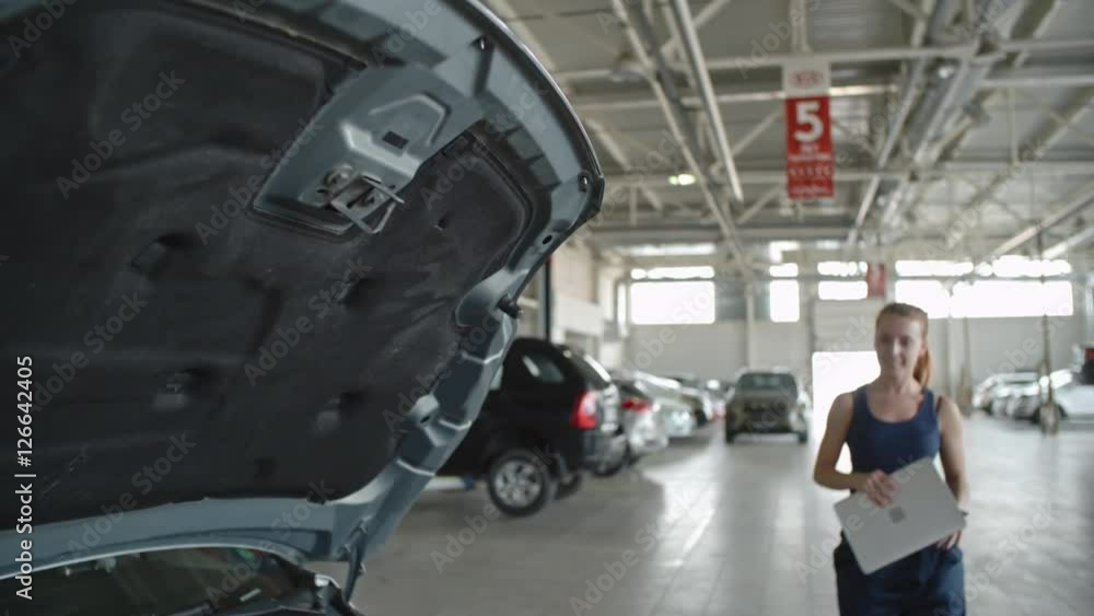 Tilt down of beautiful woman in mechanic uniform walking through repair garage and smiling while using laptop to check engine Tilt down of beautiful woman in mechanic uniform walking through repair garage and smiling while using laptop to check engine