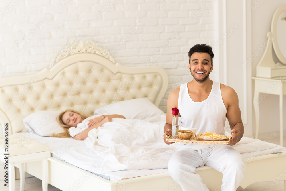 Hispanic Man Bring Breakfast To Sleeping Woman In Morning Tray With Red Rose Flower, Young Couple Lovers In Bedroom Hispanic Man Bring Breakfast To Sleeping Woman In Morning Tray With Red Rose Flower, Young Couple Lovers In Bedroom