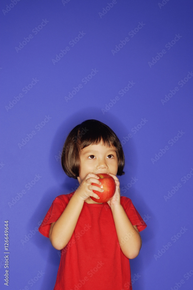 Young girl standing against blue background, eating an apple Young girl standing against blue background, eating an apple