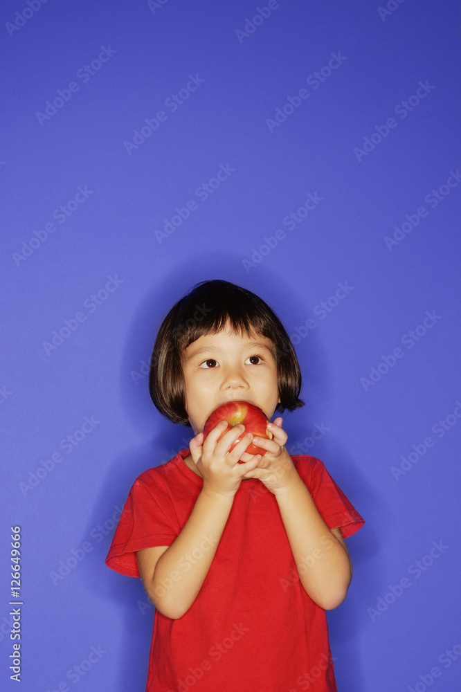 Young girl standing against blue background, biting an apple