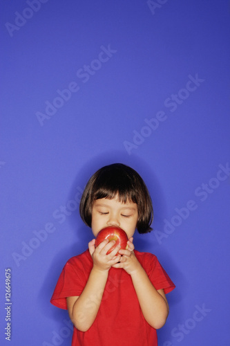 Young girl standing against blue background, holding an apple and biting it.