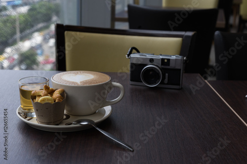 hot fresh coffee in white glass hot tea and alphabet biscuit and retro film camera with tree shape of heart foam on wooden table city view at sunset at coffee time / hot fresh coffee and tea