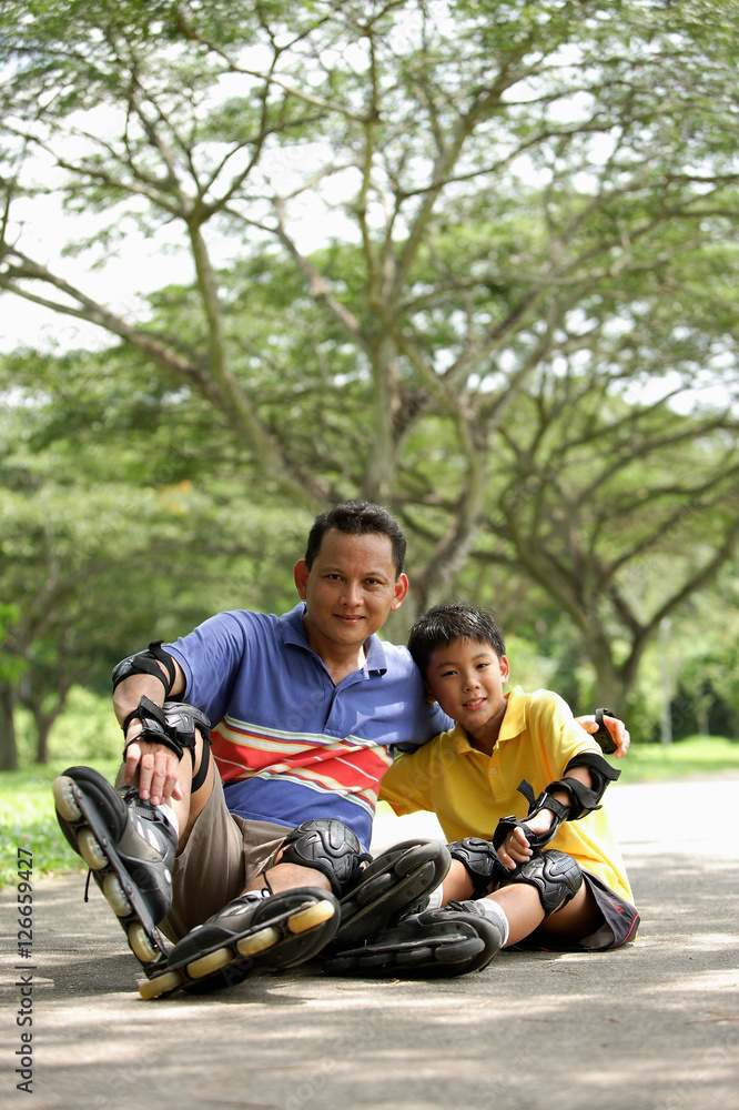 Father and son, sitting in park, wearing roller blades, looking at camera