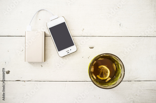 Smartphone being charged from a portable charging device, nearby there is a incomplete glass of tea with lemon