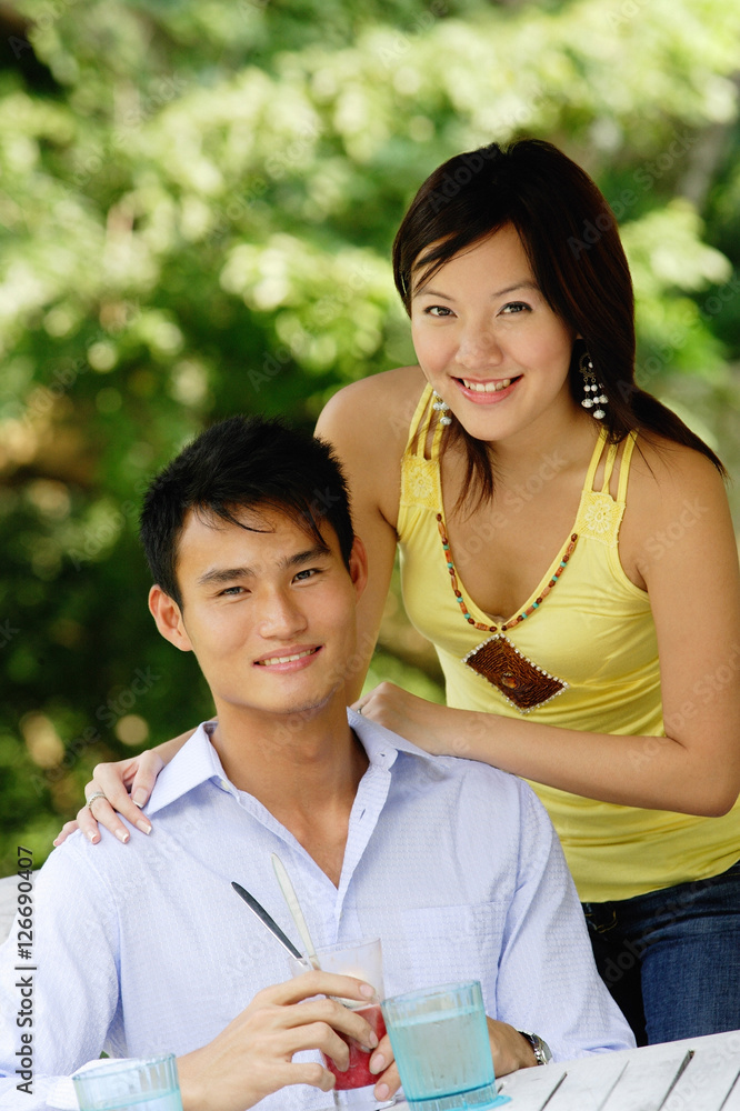 Couple smiling at camera, woman standing behind man, hand on shoulder Couple smiling at camera, woman standing behind man, hand on shoulder