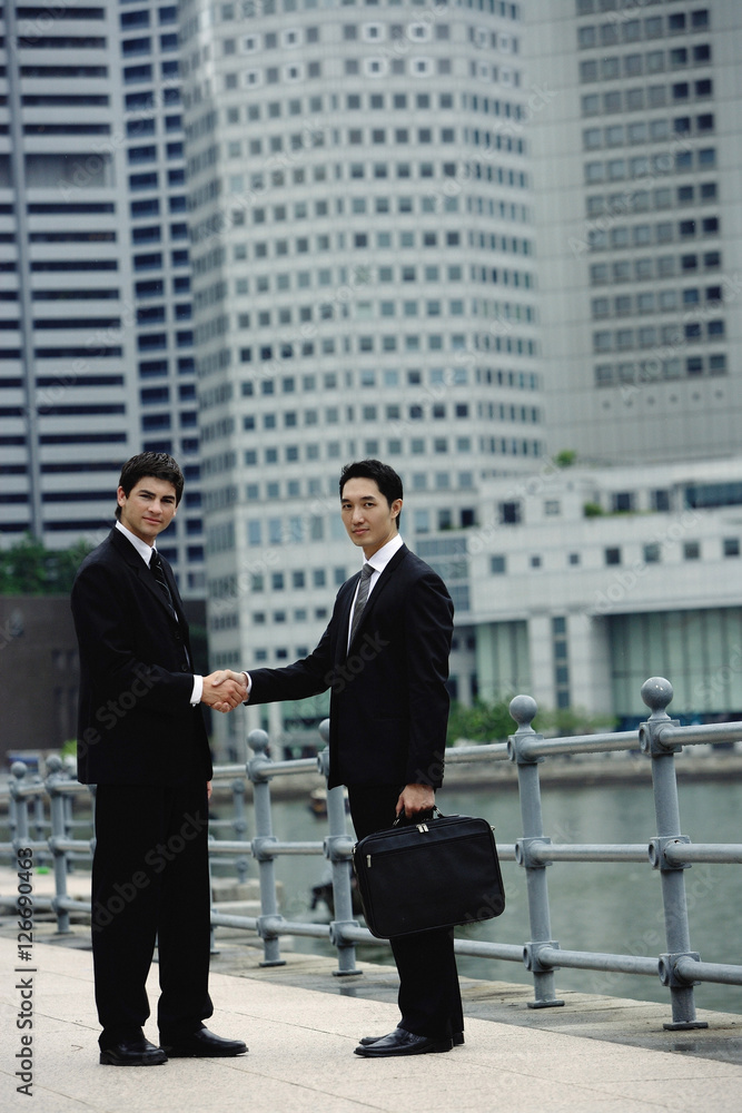 Two businessmen shaking hands, looking at camera, river and buildings in the background Two businessmen shaking hands, looking at camera, river and buildings in the background
