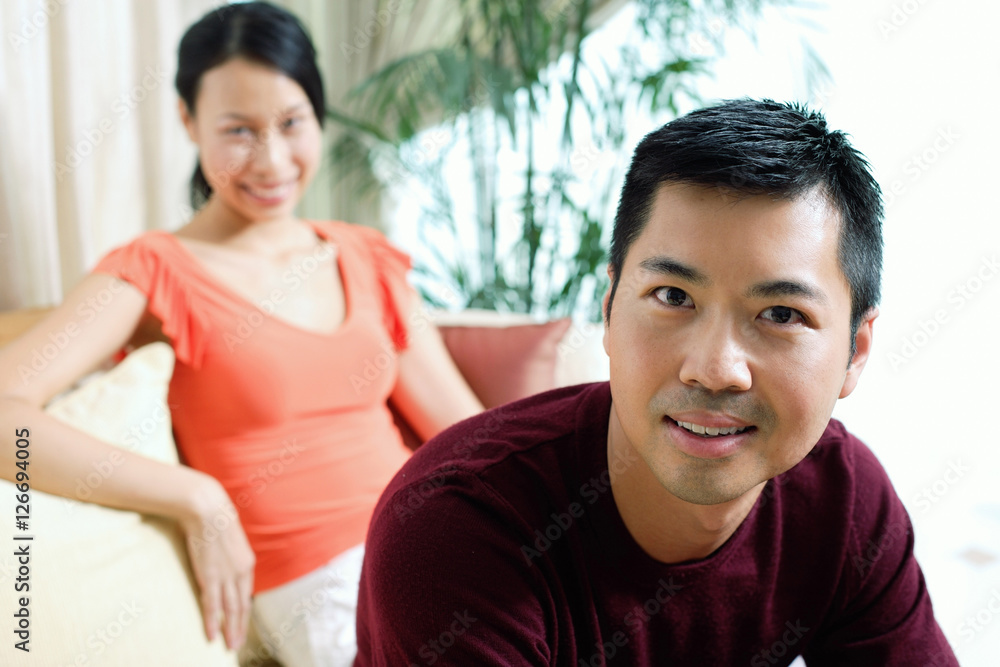 Couple at home, man looking at camera, smiling, woman sitting in the background Couple at home, man looking at camera, smiling, woman sitting in the background