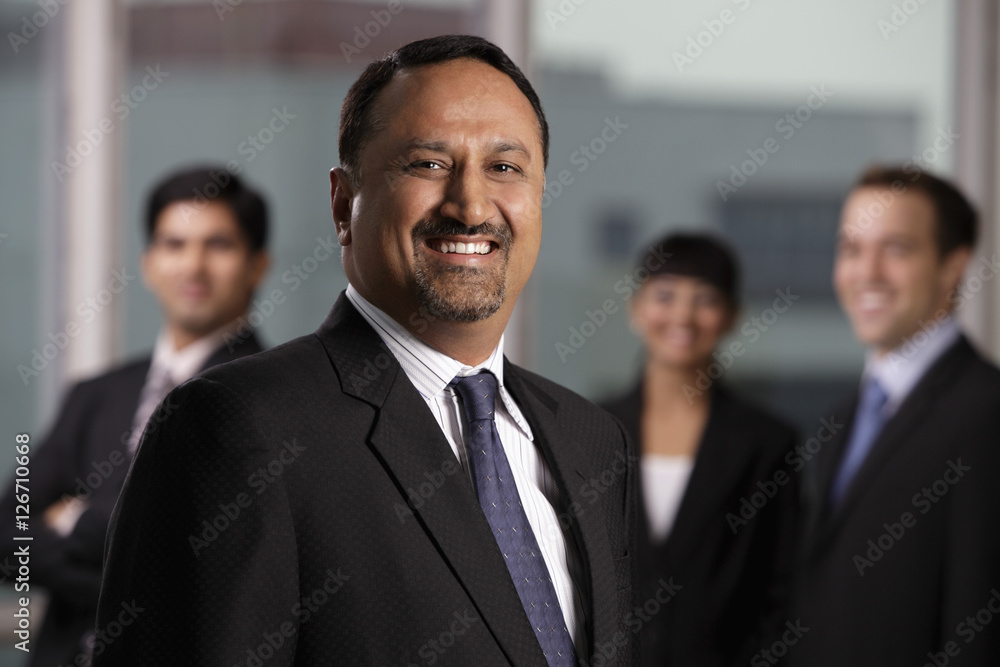 Indian man smiling at camera with colleagues behind him Indian man smiling at camera with colleagues behind him