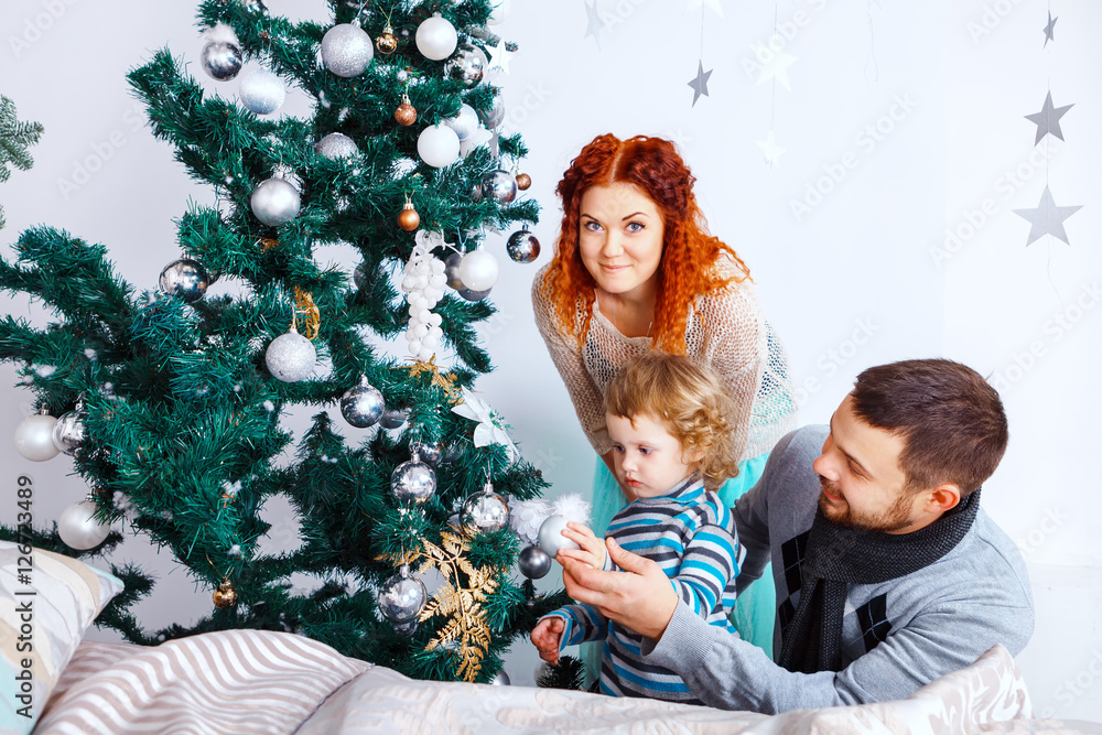 Christmas happy family of three persons decorating fir tree with gift boxes over white bedroom background Christmas happy family of three persons decorating fir tree with gift boxes over white bedroom background