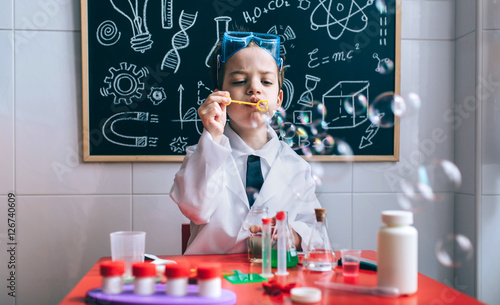 Little boy scientist playing with soap bubbles over table against of drawn blackboard