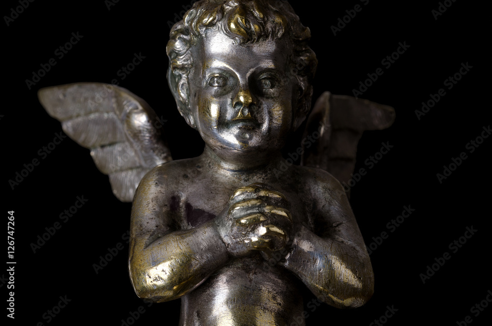 Praying winged putto on black background. Angel made of brass, covered with silver, as part of a candelabra from nineteenth century and symbol for religious passion. Macro object photo front view.