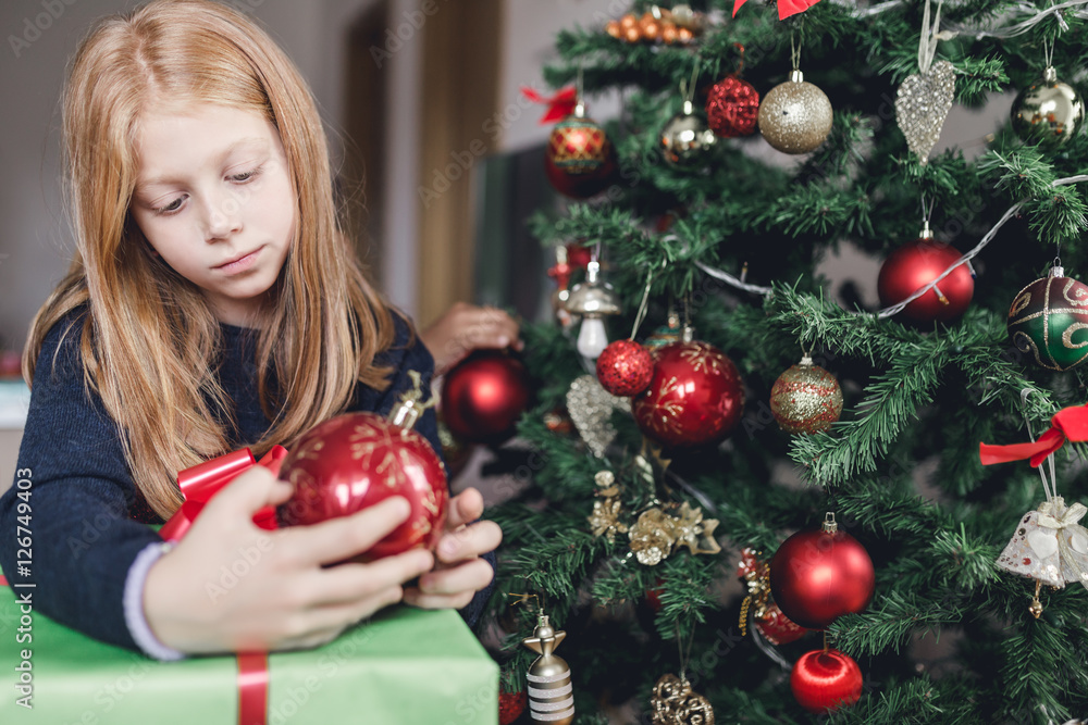 Beautiful redhead little girl holding Christmas ornament ball next to the Christmas tree. Natural light. 