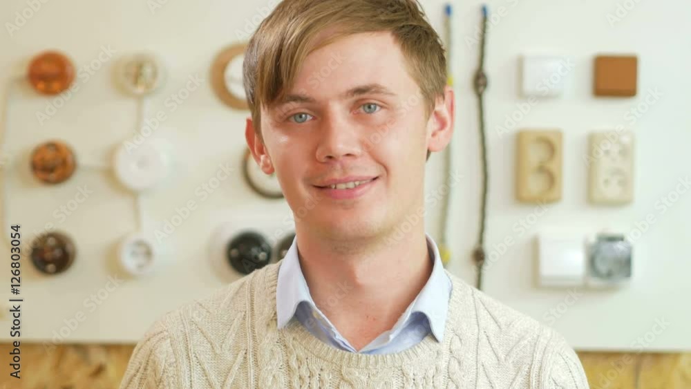 A young man smiling and looking at camera on the background of sockets and switches. Seller is dressed in a shirt and has a little stubble A young man smiling and looking at camera on the background of sockets and switches. Seller is dressed in a shirt and has a little stubble