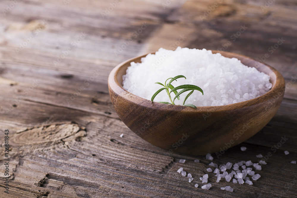 Large White sea salt in a natural wooden bowl with sprig of fresh rosemary on simple background. Selective focus. Large White sea salt in a natural wooden bowl with sprig of fresh rosemary on simple background. Selective focus.