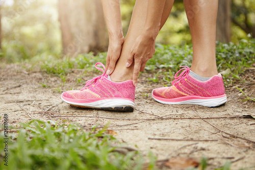 Woman runner holding her twisted ankle after running exercise outdoors. Female jogger in pink sneakers clutching calf muscle after spraining it while out jogging on trail in summer outdoors nature