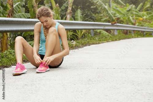 Young tired Caucasian woman runner lacing her pink running shoes, sitting on road in tropical forest having small break while jogging outdoors. Female jogger tying laces on her sneakers during workout