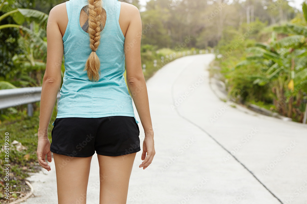 Back view of blonde woman runner with beautiful body dressed in sportswear standing on road in tropical forest before morning training. Half length view from behind with copy space. Selective focus Back view of blonde woman runner with beautiful body dressed in sportswear standing on road in tropical forest before morning training. Half length view from behind with copy space. Selective focus