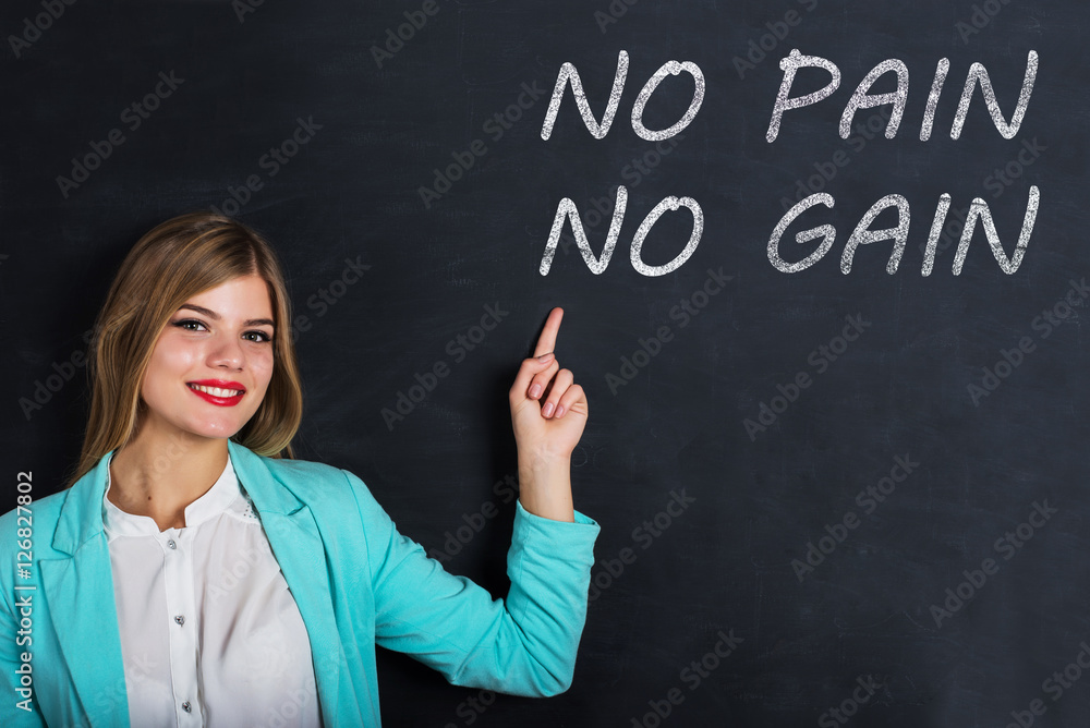Beautiful young female student is pointing, looking at camera and smiling, standing against blackboard with text Beautiful young female student is pointing, looking at camera and smiling, standing against blackboard with text