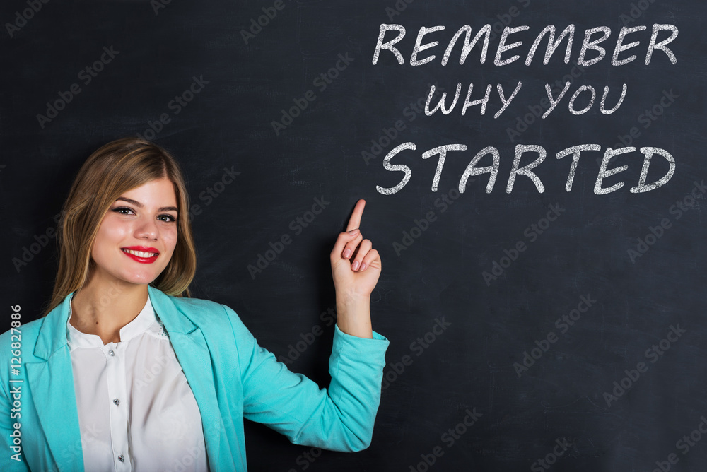Beautiful young female student is pointing, looking at camera and smiling, standing against blackboard with text Beautiful young female student is pointing, looking at camera and smiling, standing against blackboard with text