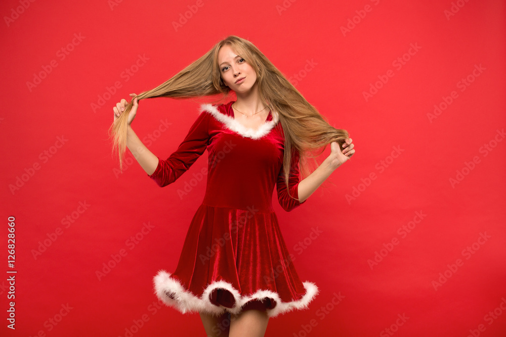 Beautiful young woman in Santa costume playing with a strand of her long silky hair, looking at camera on red background Beautiful young woman in Santa costume playing with a strand of her long silky hair, looking at camera on red background