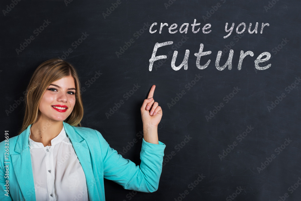 Beautiful young female student is pointing, looking at camera and smiling, standing against blackboard with text Beautiful young female student is pointing, looking at camera and smiling, standing against blackboard with text
