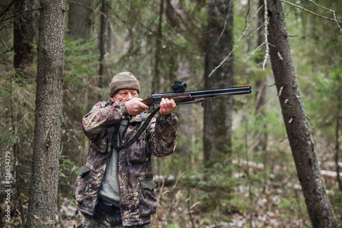 Male hunter shoots a gun. Hunter wearing a camouflage jacket and cap. A man of 50 years. On the ground lies a little of snow. Cloudy weather, autumn.