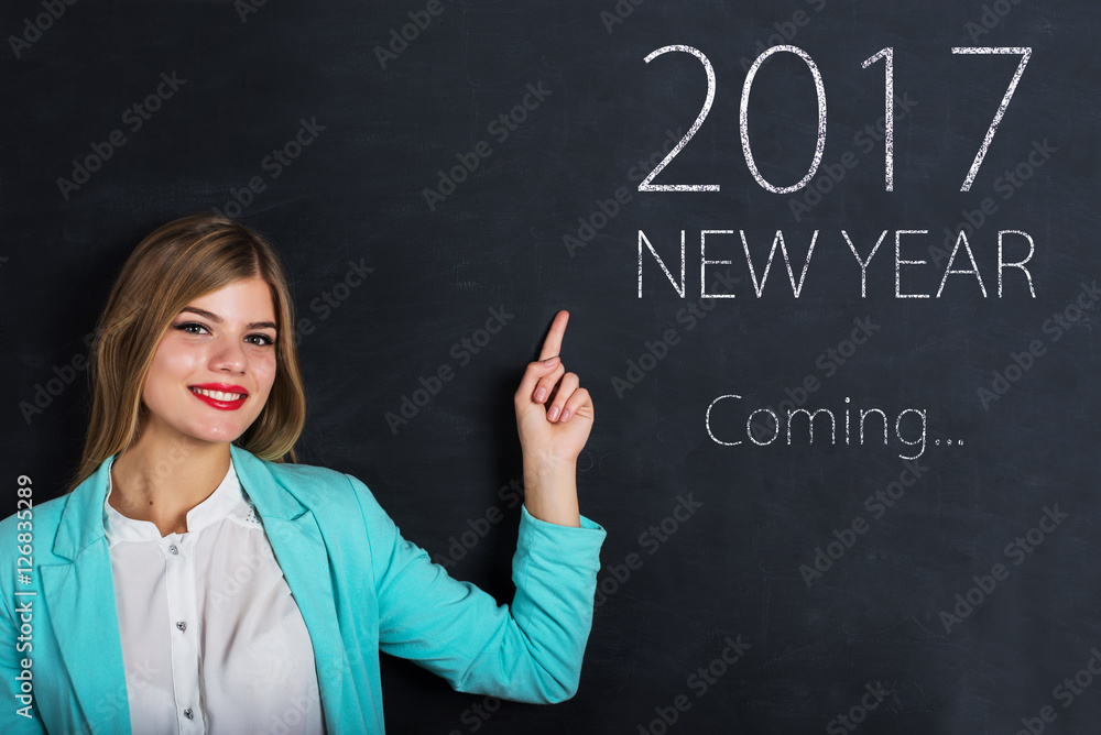 Beautiful young female student is pointing, looking at camera and smiling, standing against blackboard with text Beautiful young female student is pointing, looking at camera and smiling, standing against blackboard with text