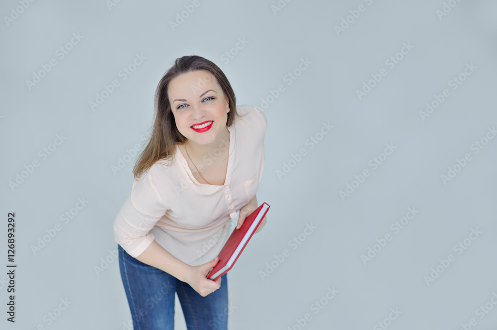 Young woman holding a red book and smiles broadly, leaning forward, grey background, copy space. Young woman holding a red book and smiles broadly, leaning forward, grey background, copy space.